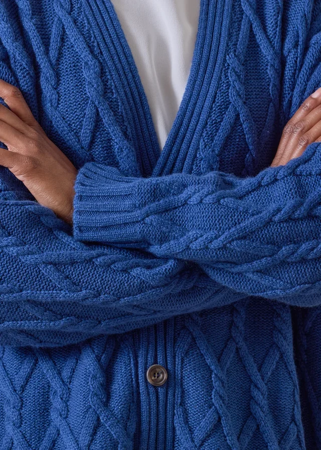 Blue cable cardigan with ribbed cuffs and front edging, shown on a model with folded arms.