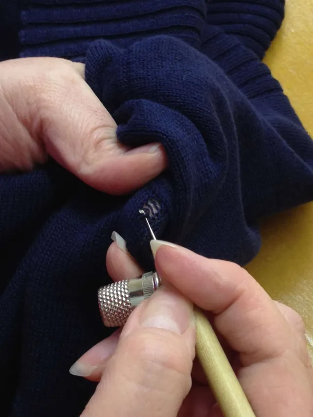 Hands repairing a hole in navy knitted fabric with a fine tool.